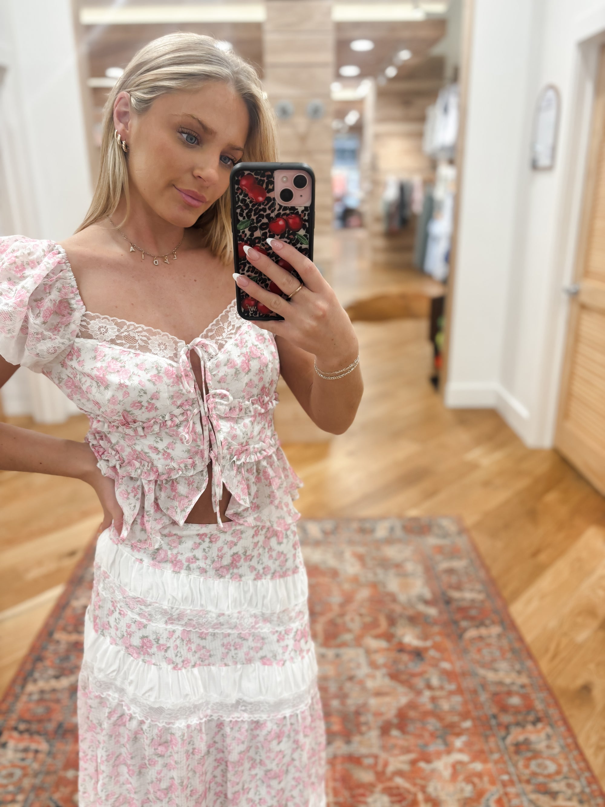 Sweet and feminine, this tiered maxi skirt showcases a soft pink and white ditsy rose print with delicate lace inset detailing. The flowing silhouette and romantic layers create a vintage-inspired look that’s perfect for garden parties or summer strolls. Pair it with the matching puff sleeve crop top featuring a ruched front and bow tie detail for the complete romantic set.