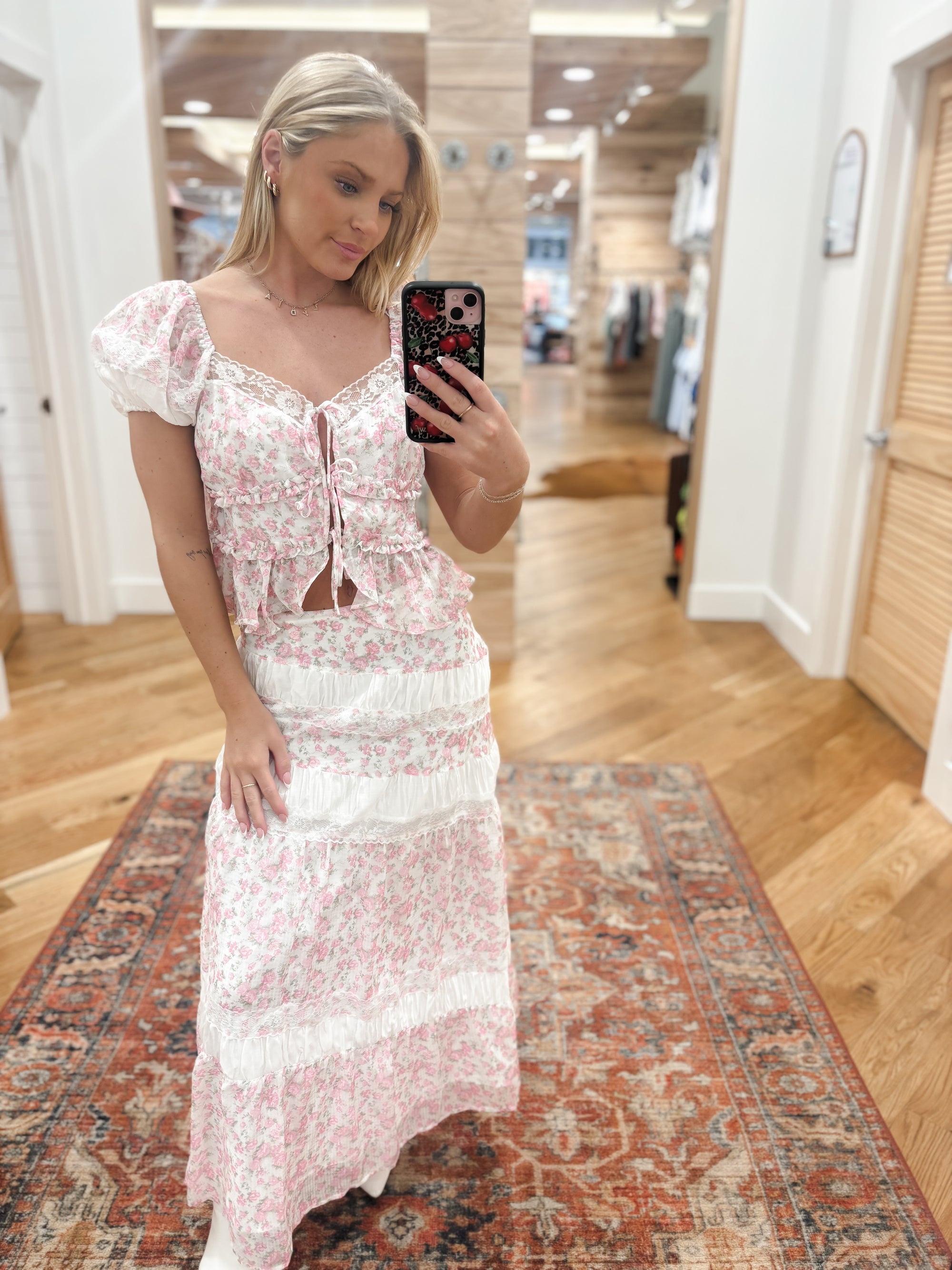 Sweet and feminine, this tiered maxi skirt showcases a soft pink and white ditsy rose print with delicate lace inset detailing. The flowing silhouette and romantic layers create a vintage-inspired look that’s perfect for garden parties or summer strolls. Pair it with the matching puff sleeve crop top featuring a ruched front and bow tie detail for the complete romantic set.